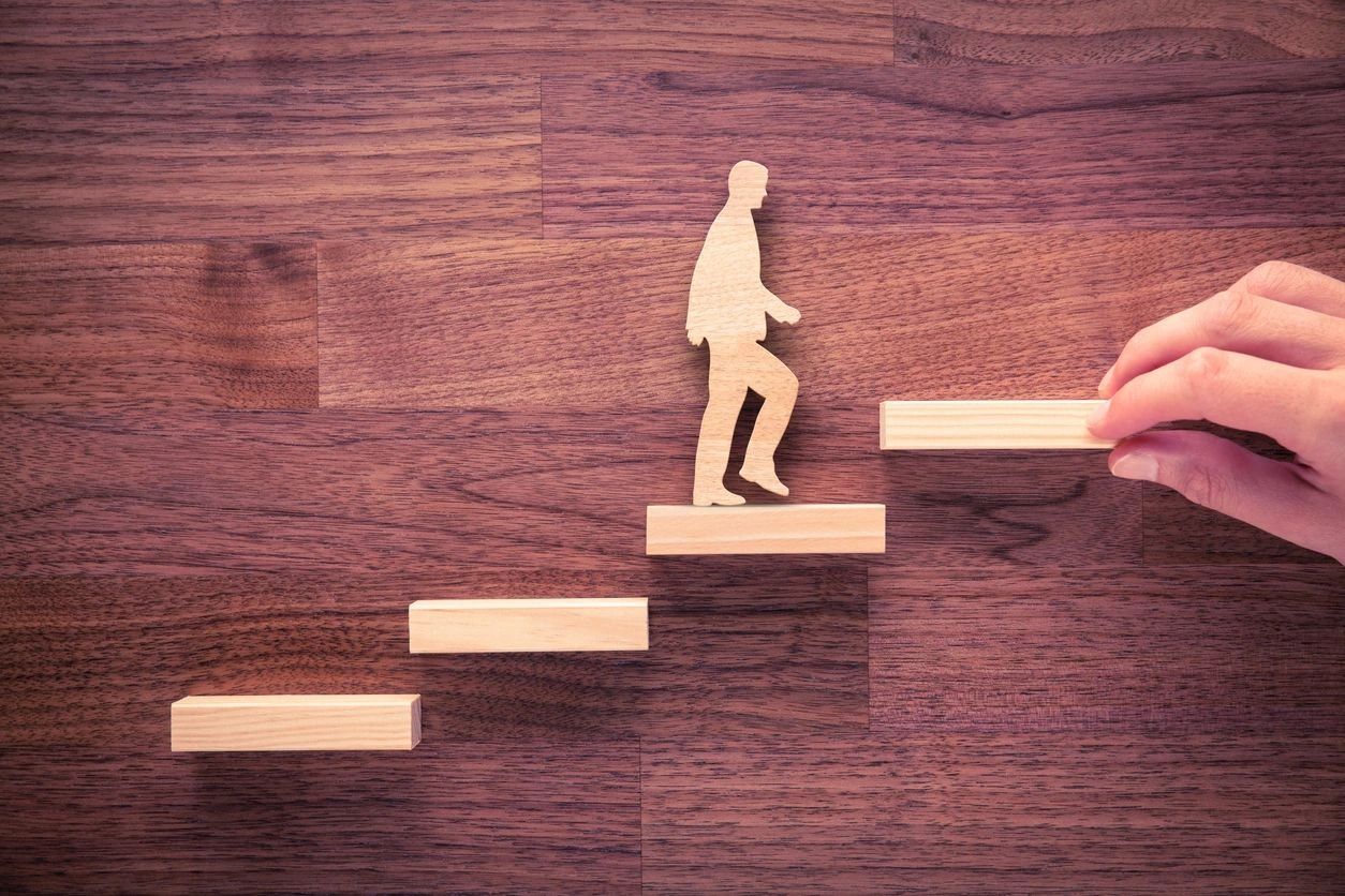 Wooden figure climbing stairs made of wood blocks, a hand placing the next step — leadership growth concept