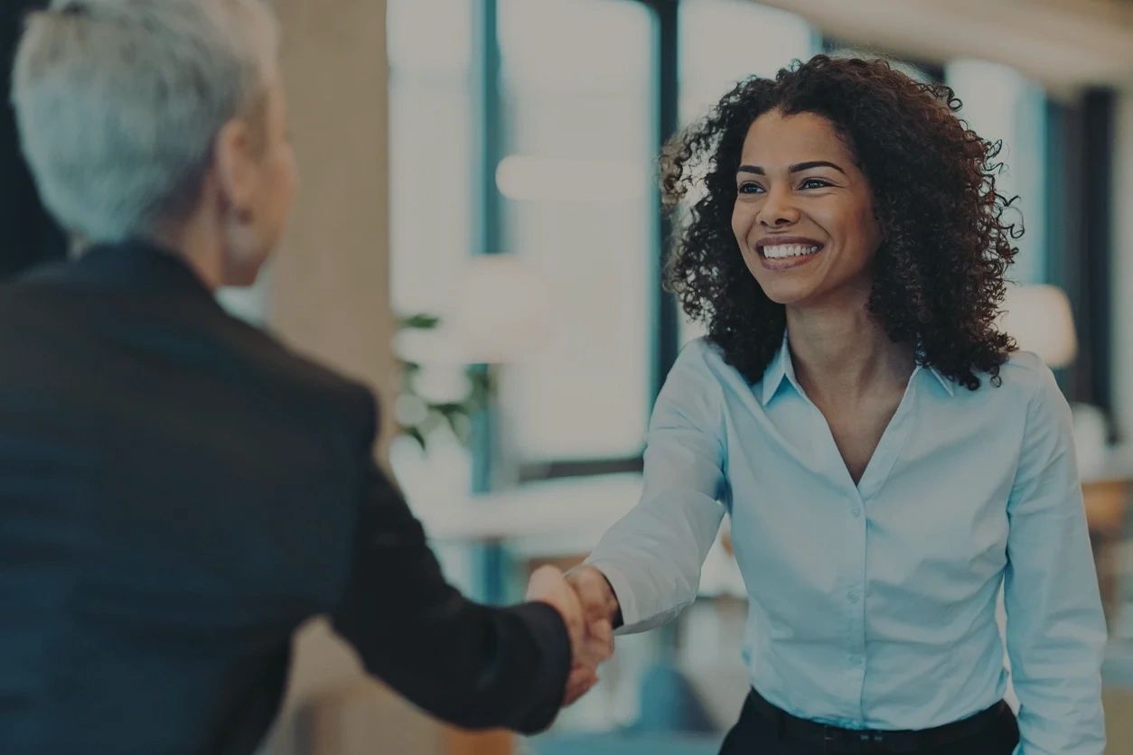 Two professionals shaking hands in a modern office, a smiling woman with curly hair in the foreground