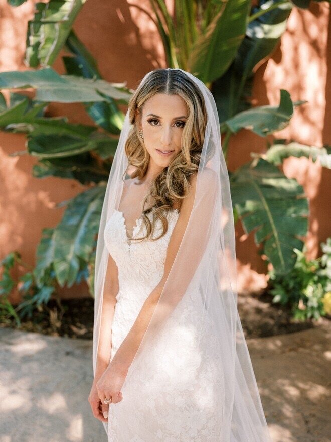 Blonde bride in lace gown and veil standing against a terracotta wall with tropical plants