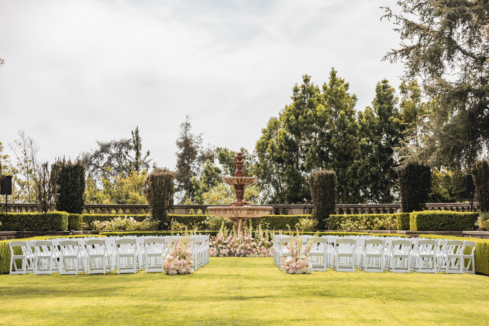 Elegant outdoor wedding ceremony on a manicured lawn with a fountain, white chairs and pink florals