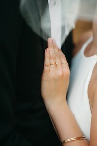 A bride's hand with engagement ring resting on the groom's chest near a white veil
