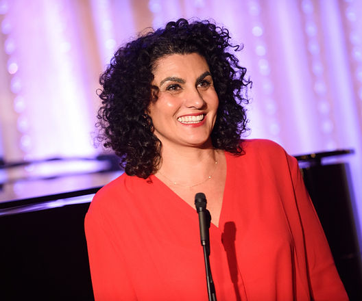 Lorenza Muñoz speaking at a microphone on a purple-lit stage in a red blouse