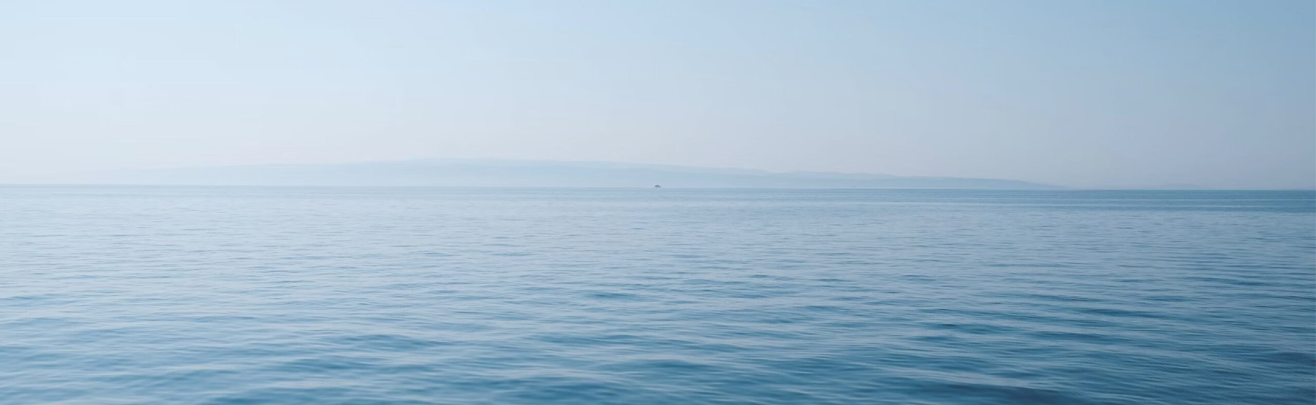 Calm open ocean meeting a hazy horizon under a pale blue sky