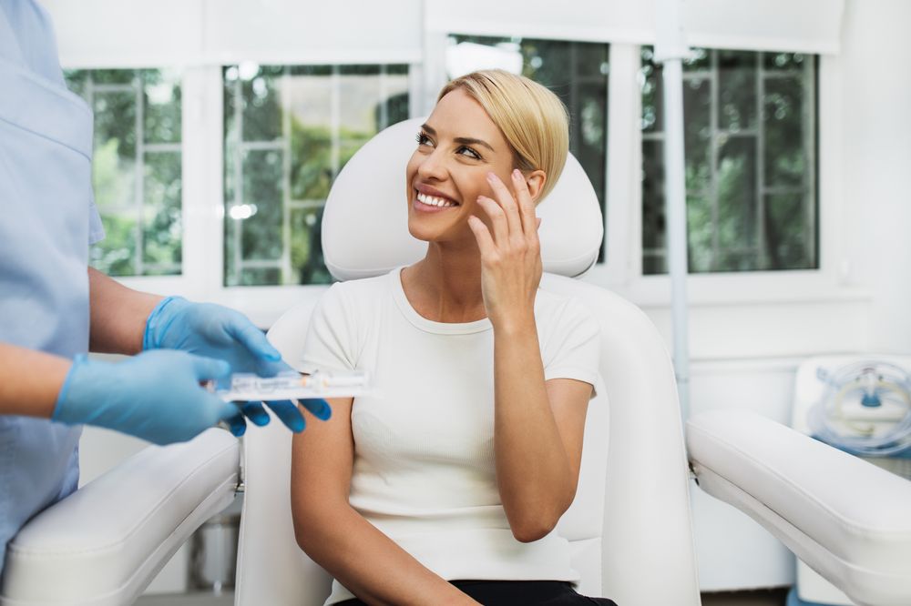 Smiling client seated in a white treatment chair while a practitioner in blue gloves performs an aesthetic treatment