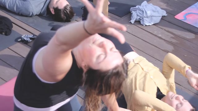 People practicing yoga on a wooden rooftop deck in the East Village, a woman in the foreground gently arching into a backbend in warm sunlight