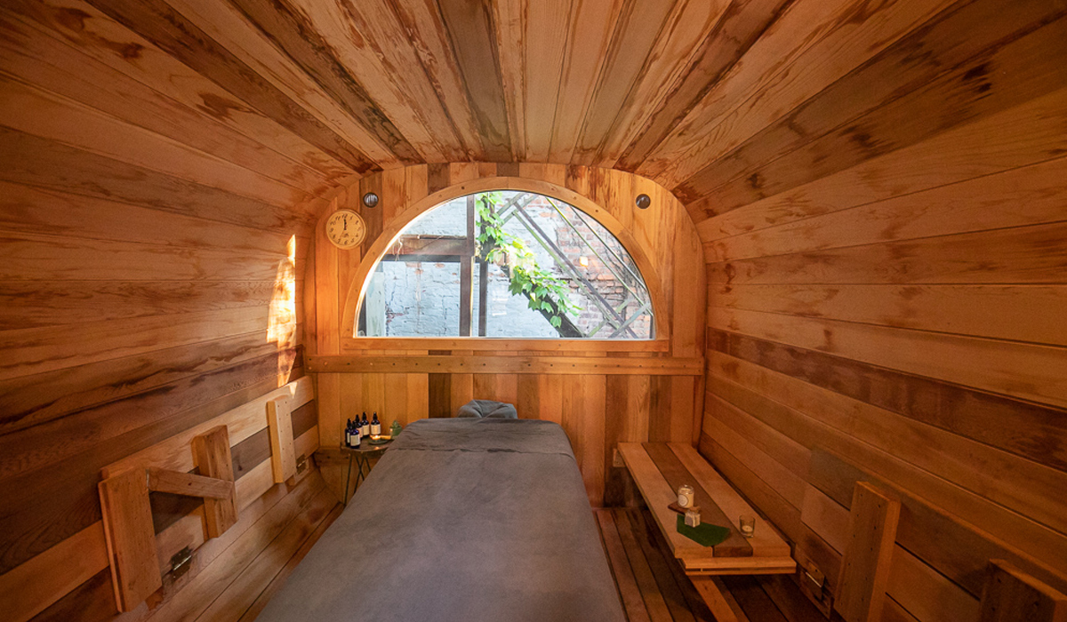 Barrel-shaped cedar sauna treatment room interior with an arched window and massage table