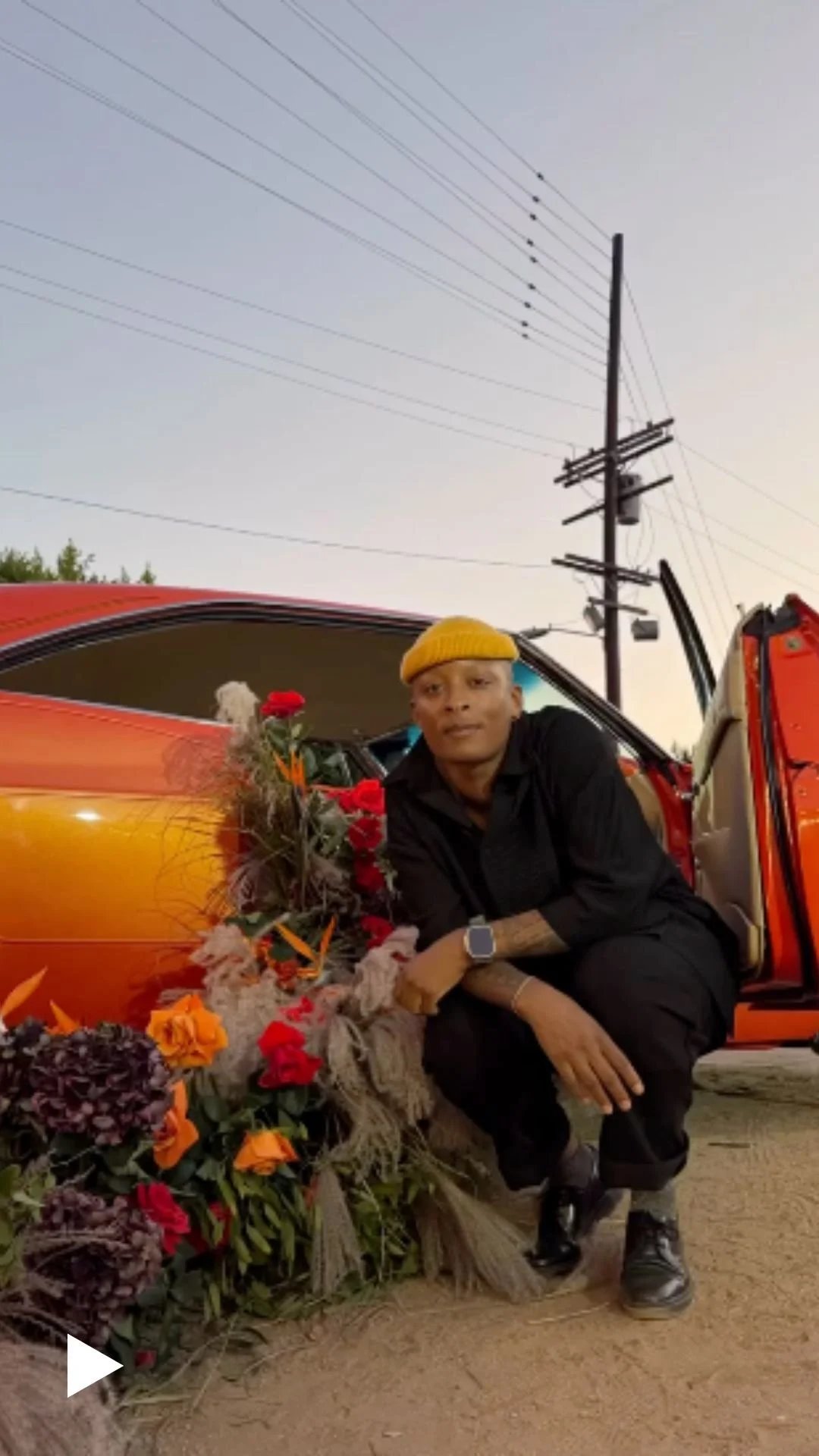 Person in a yellow beanie beside a floral arrangement next to a vintage car