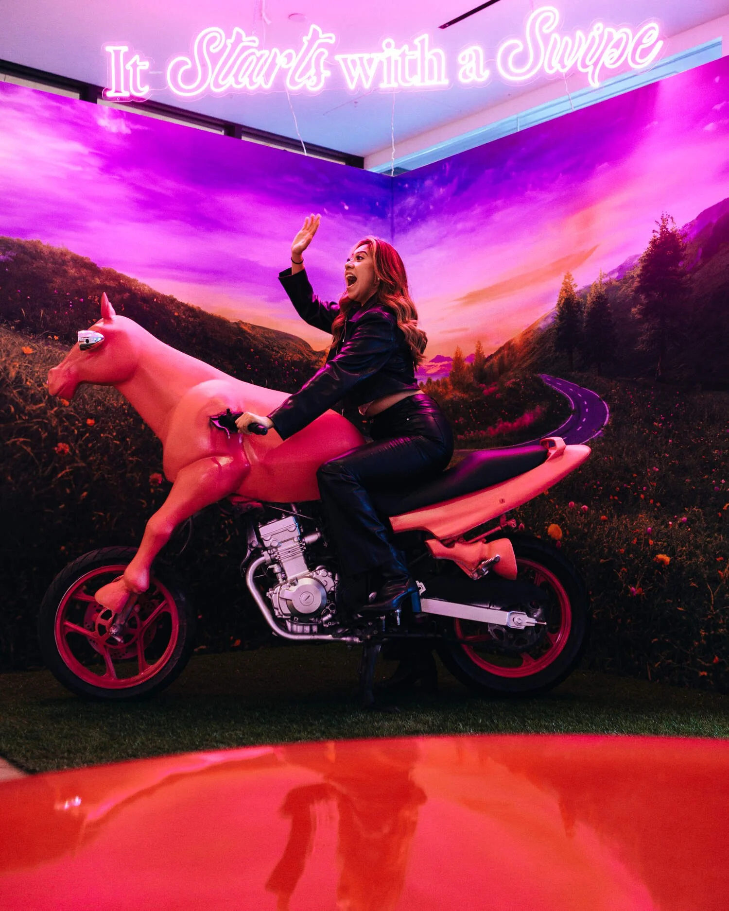 Woman riding a pink horse-motorcycle in a neon Tinder installation reading It Starts with a Swipe