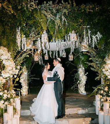 Bride and groom under a floral chuppah with candles and lanterns at a night wedding