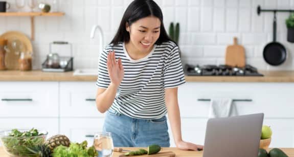 Woman waving at her laptop in a bright kitchen with vegetables on the counter