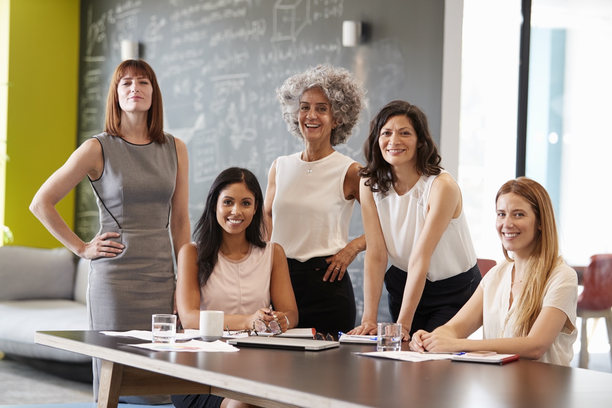 Five professional women smiling around a conference table in a modern office