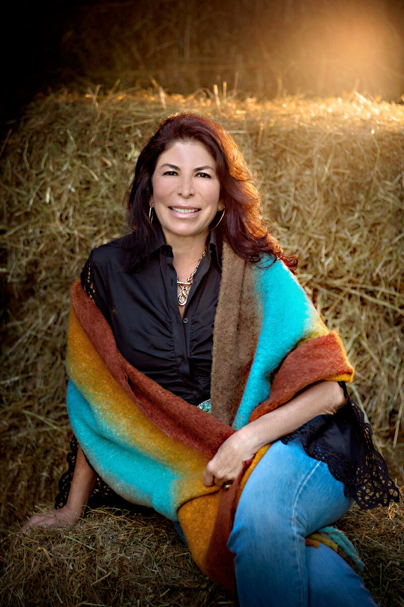 Lisa Cypers Kamen in a colorful shawl sitting against hay bales, smiling warmly