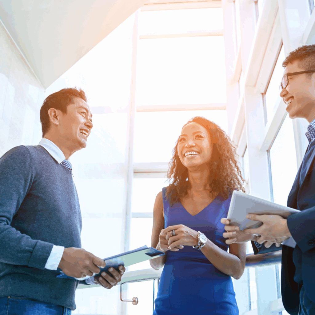 Three smiling business professionals having a casual conversation in a bright, light-flooded office
