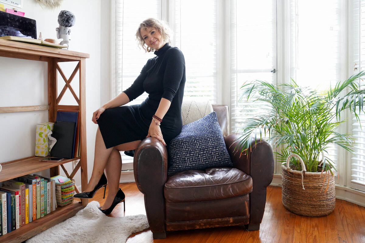 Jacqueline in a black turtleneck dress on a leather armchair in her bright home office