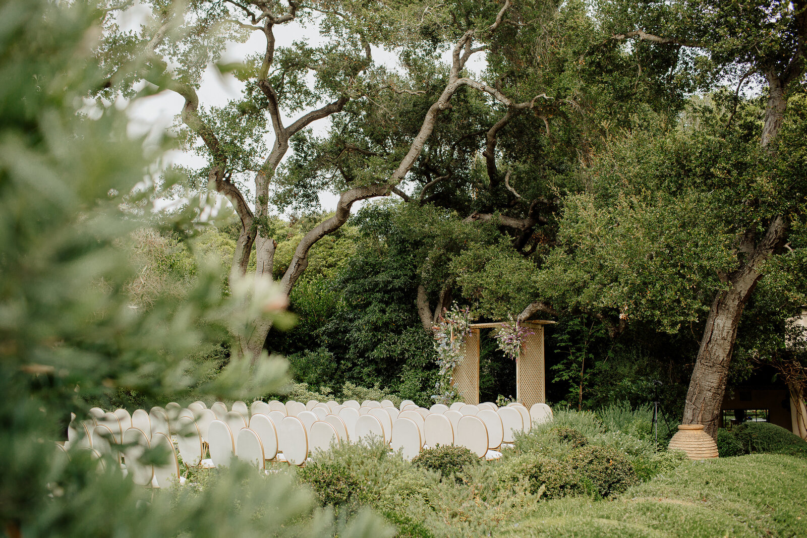 Outdoor wedding ceremony with white chairs and a floral arch set beneath grand oak trees
