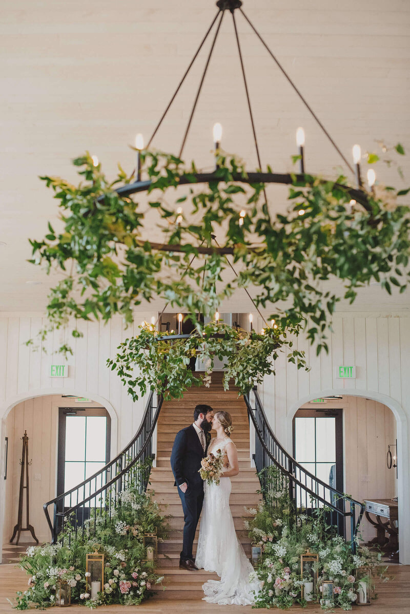Couple kissing at the base of a grand barn staircase decorated with a greenery chandelier — Wes and Victoria