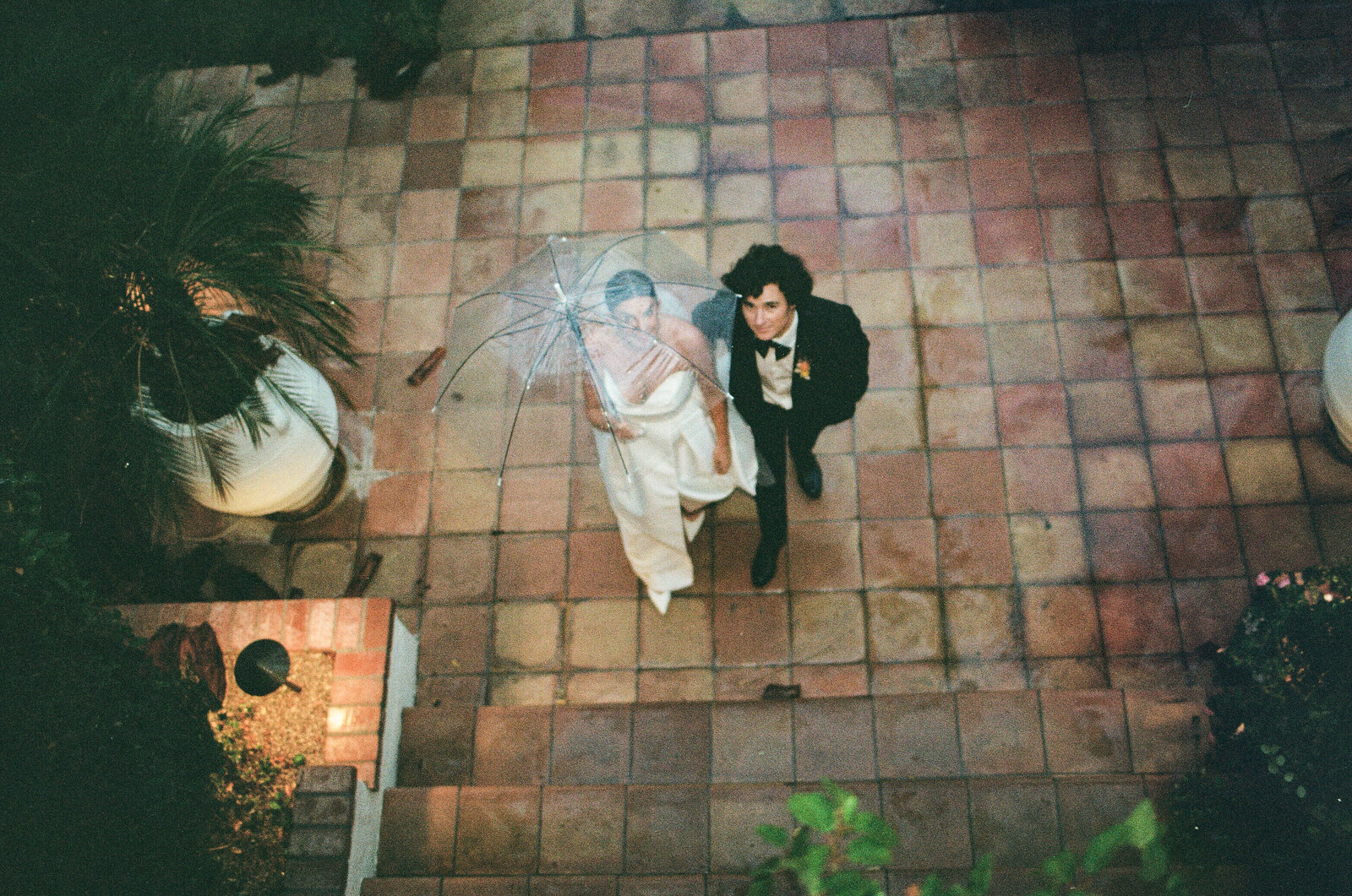 Aerial view of a bride and groom on a terracotta tile patio with a clear umbrella