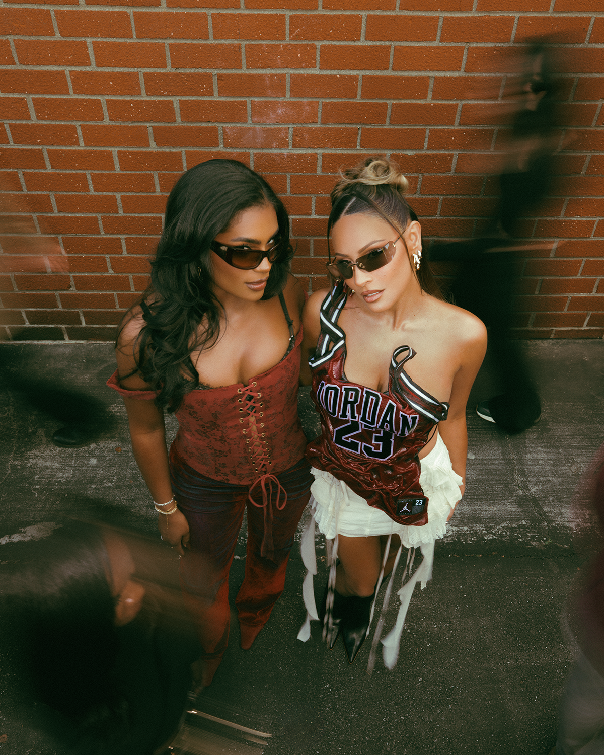 Desi Perkins and Monet McMichael wearing Dezi Eyewear sunglasses against a brick wall.