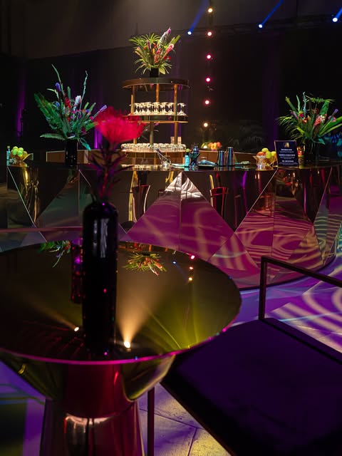 Moody nighttime event interior with reflective tables, tropical florals and stage lighting
