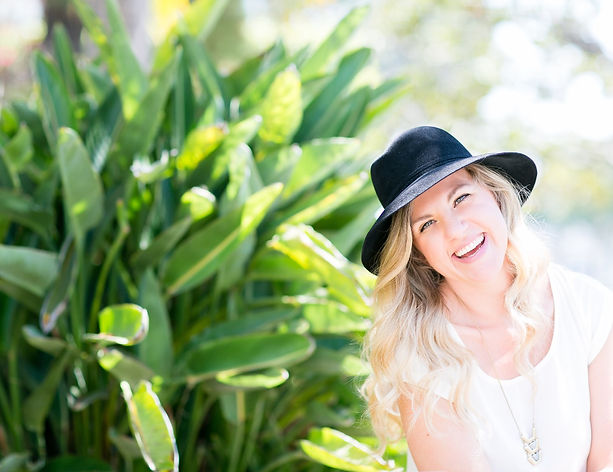 Natasha Uspensky, smiling, wearing a black hat outdoors in front of tropical foliage