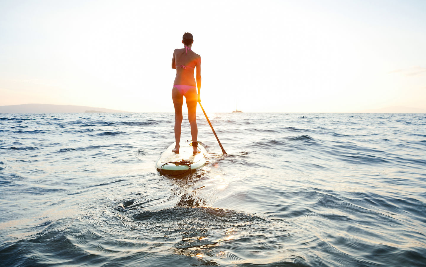 Woman on a paddleboard on calm ocean water at sunset, viewed from behind