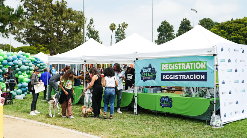 Outdoor community event with white pop-up tents, a 'Take Action' registration booth, attendees and dogs on grass