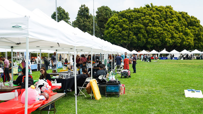 Row of white pop-up tents and attendees on a large grass field with trees
