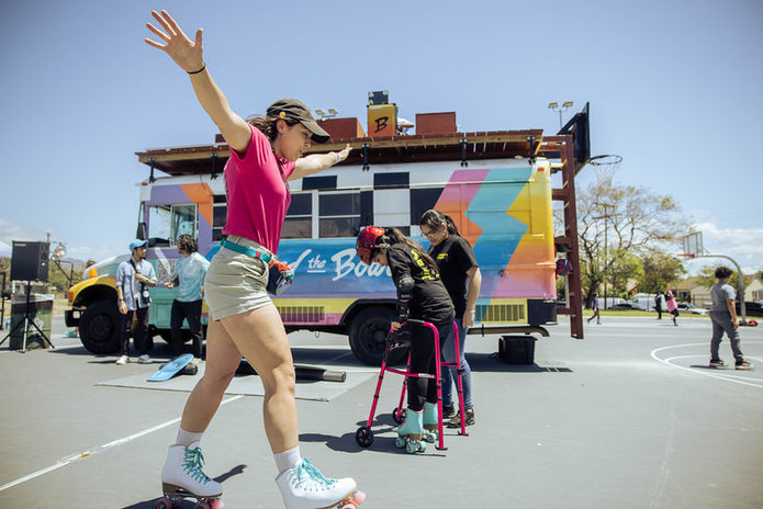 Woman on roller skates with arm raised in front of a colorful 'Bowl' branded bus at an outdoor PDSC community event on a basketball court