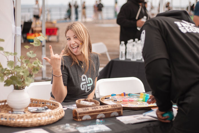 Smiling blonde woman in a branded t-shirt at an event vendor table by the pier