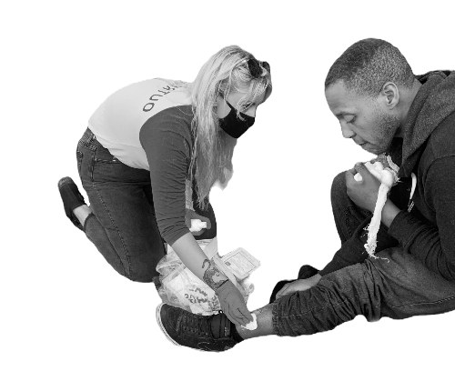 Black-and-white photo of a woman kneeling to provide wound care to a man seated on the ground.