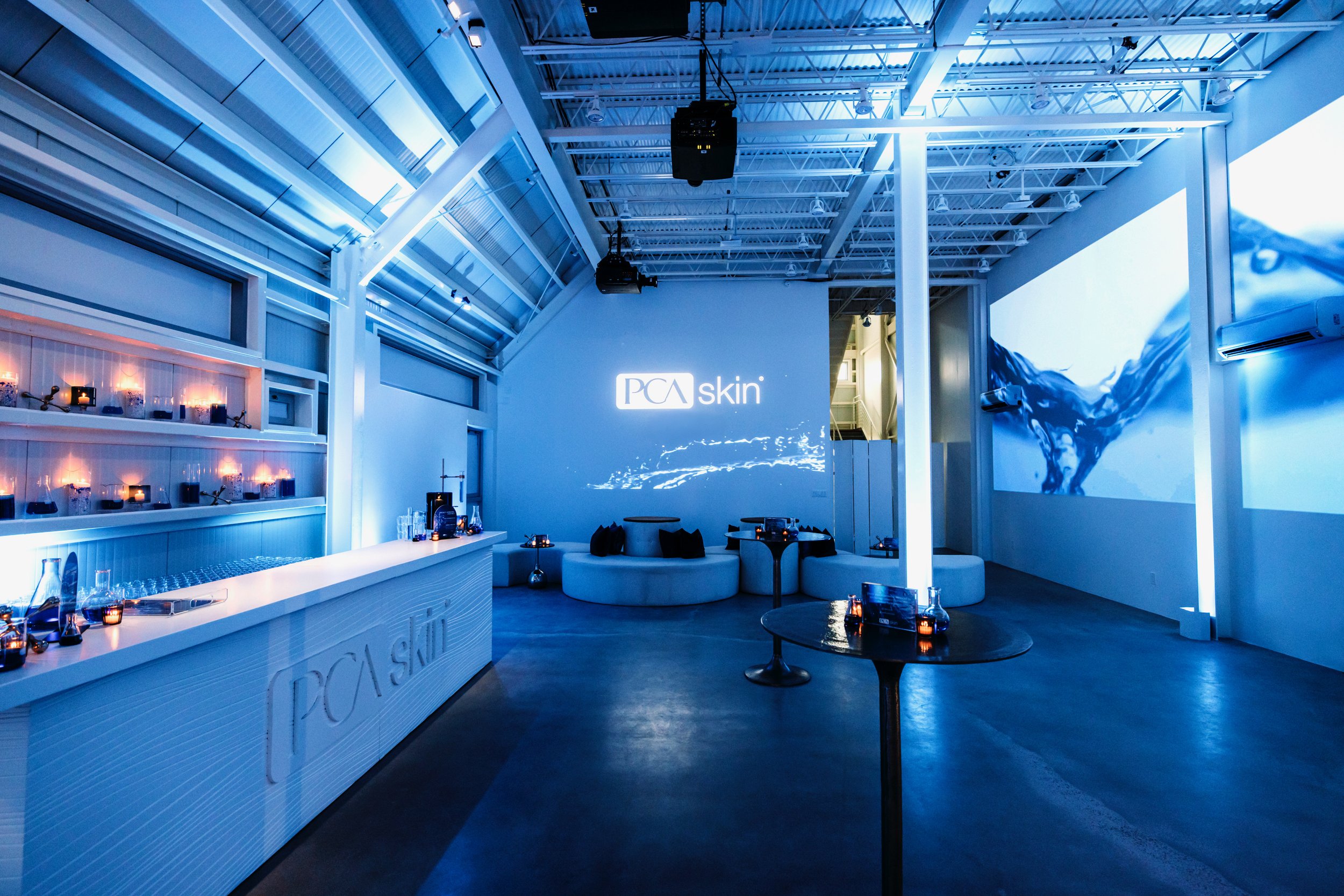 PCA Skin event space with blue lighting, branded bar counter, lounge seating and large water projection screen