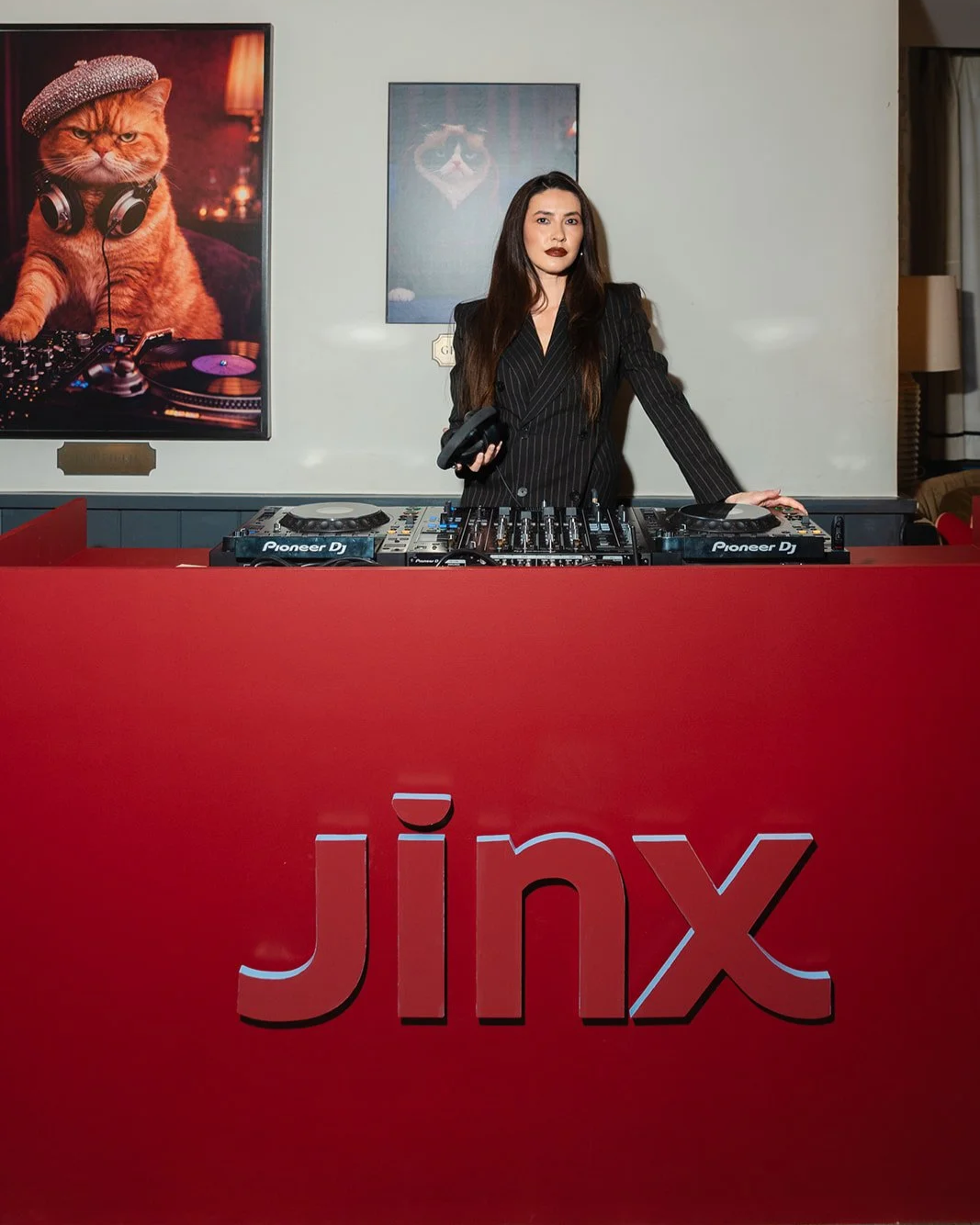 Female DJ in a black pinstripe blazer at a red Jinx-branded DJ booth with cat art prints on wall behind