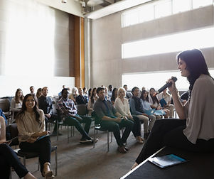 A woman speaking at a podium with a microphone to a seated seminar audience.