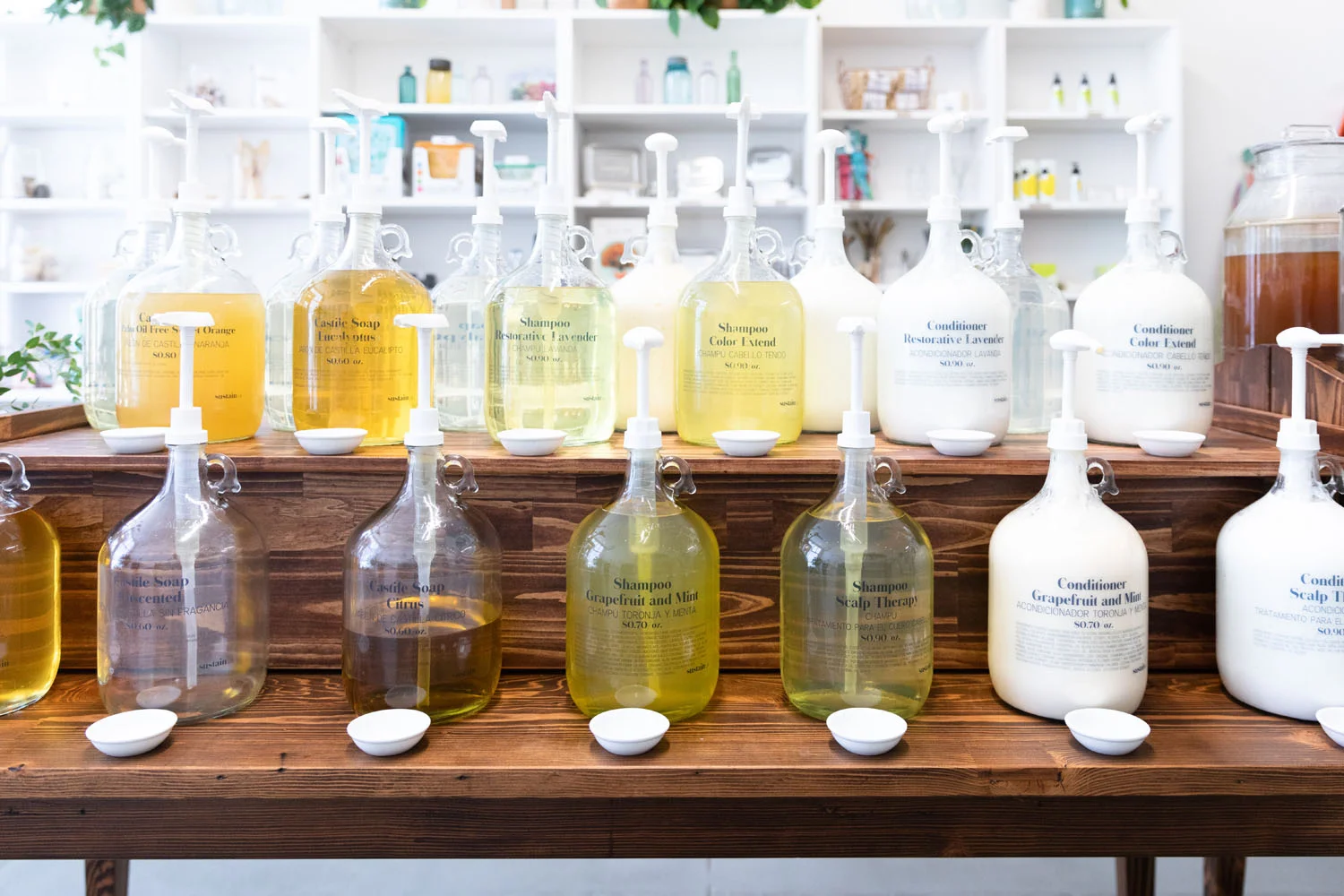 Glass refill jugs of shampoo, conditioner and castile soap on the Sustain LA refill station wooden shelving