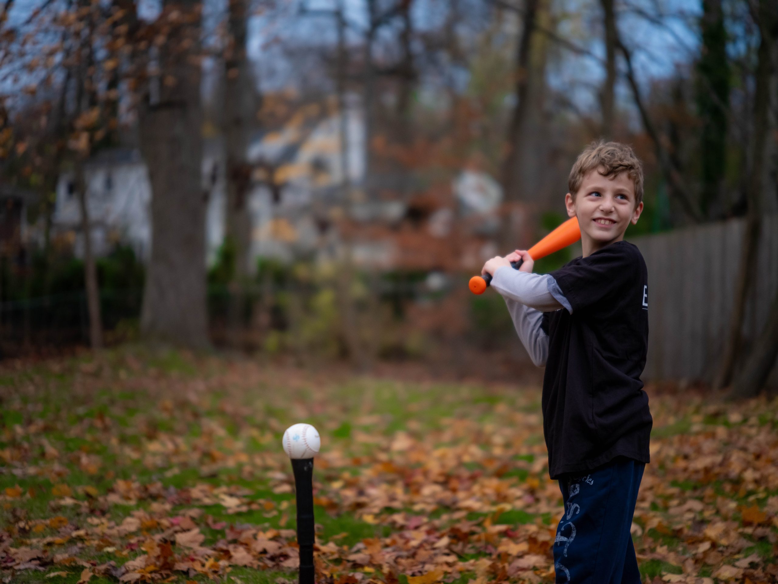Young boy smiling while swinging an orange bat at a tee in an autumn backyard
