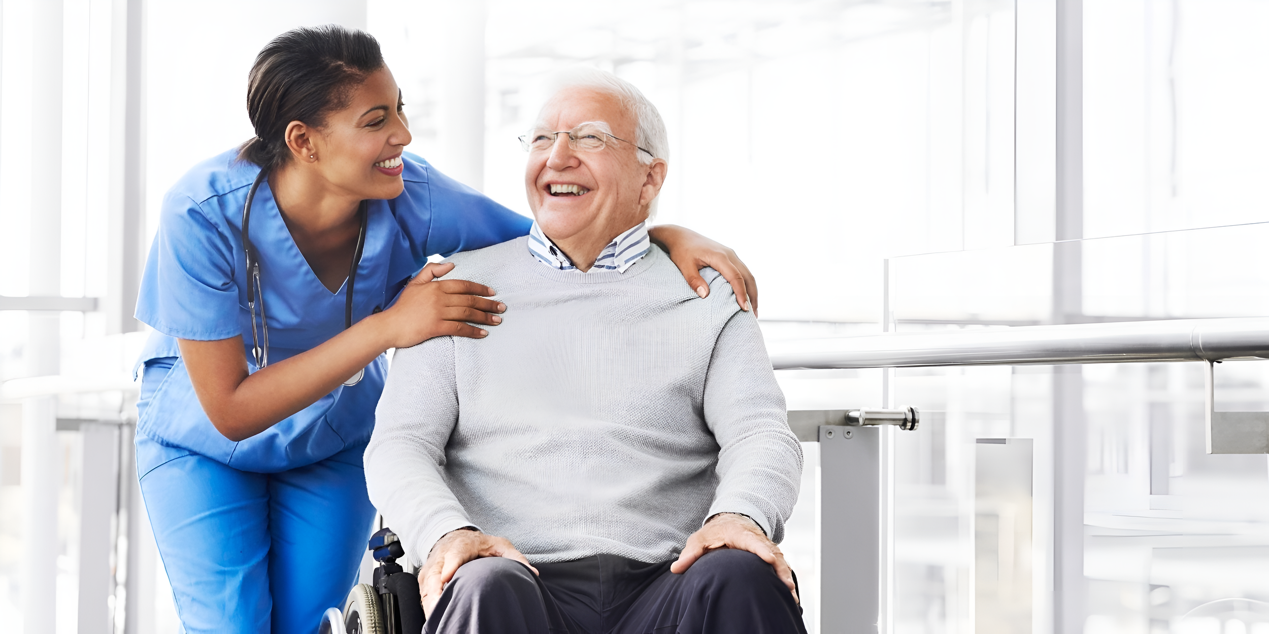 A nurse in blue scrubs smiling warmly with an elderly man in a wheelchair in a bright clinical hallway