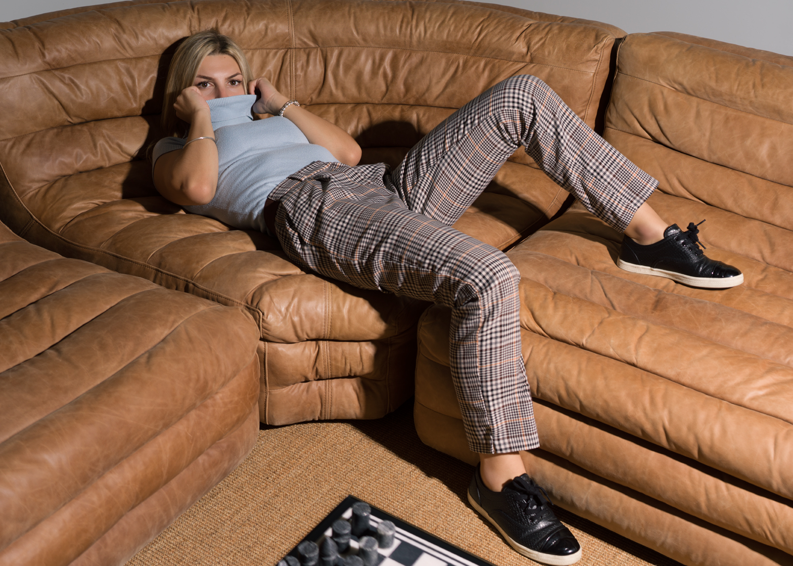 Antonia Turilli reclining on a tan leather sofa in a light-blue turtleneck and plaid trousers