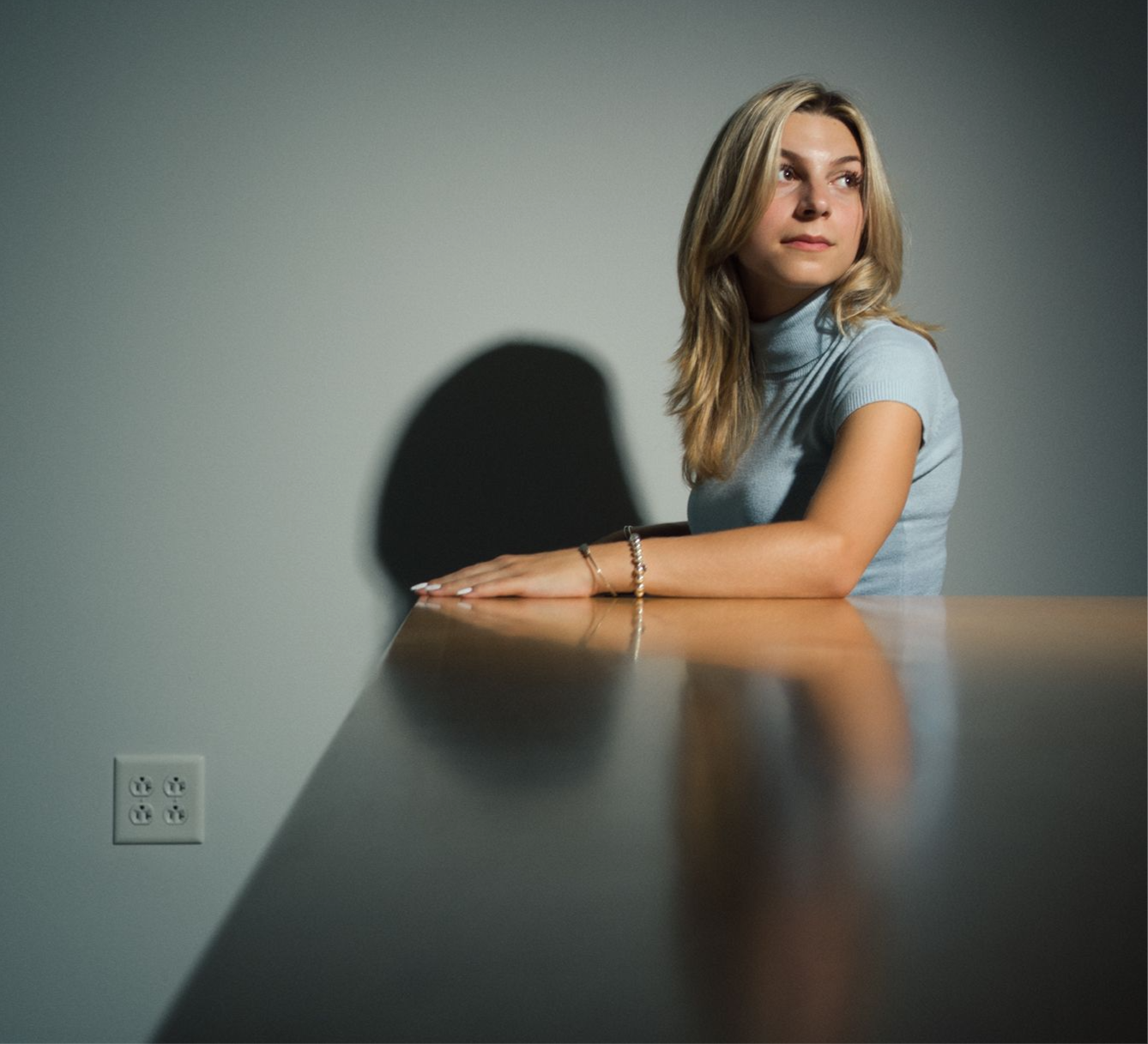 Antonia Turilli seated at a dark table, arms folded forward, dramatic shadow on a grey wall, wearing a light-blue turtleneck
