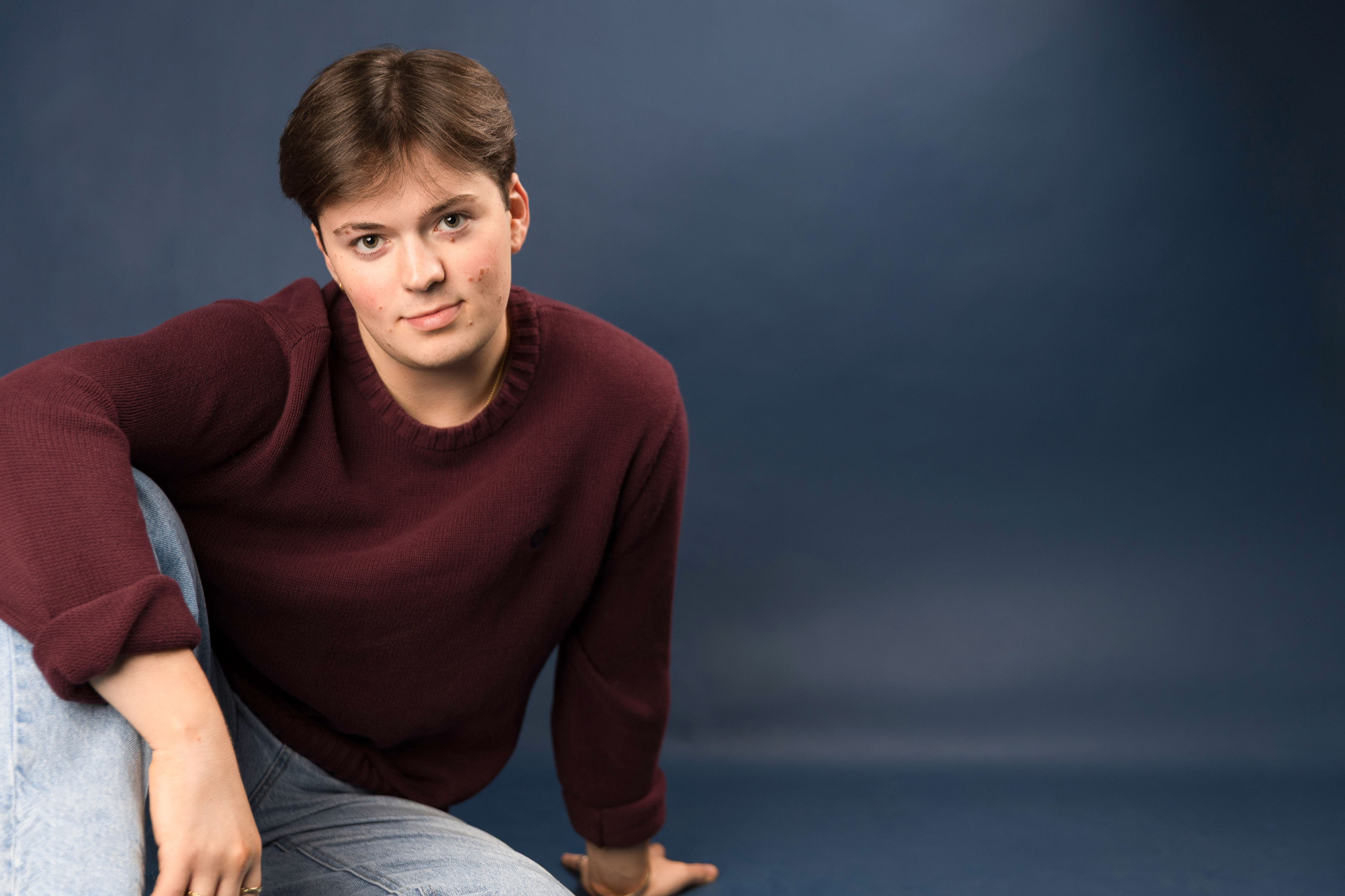 Benji Godley-Fisher seated in a burgundy sweater, leaning forward against a dark blue studio backdrop