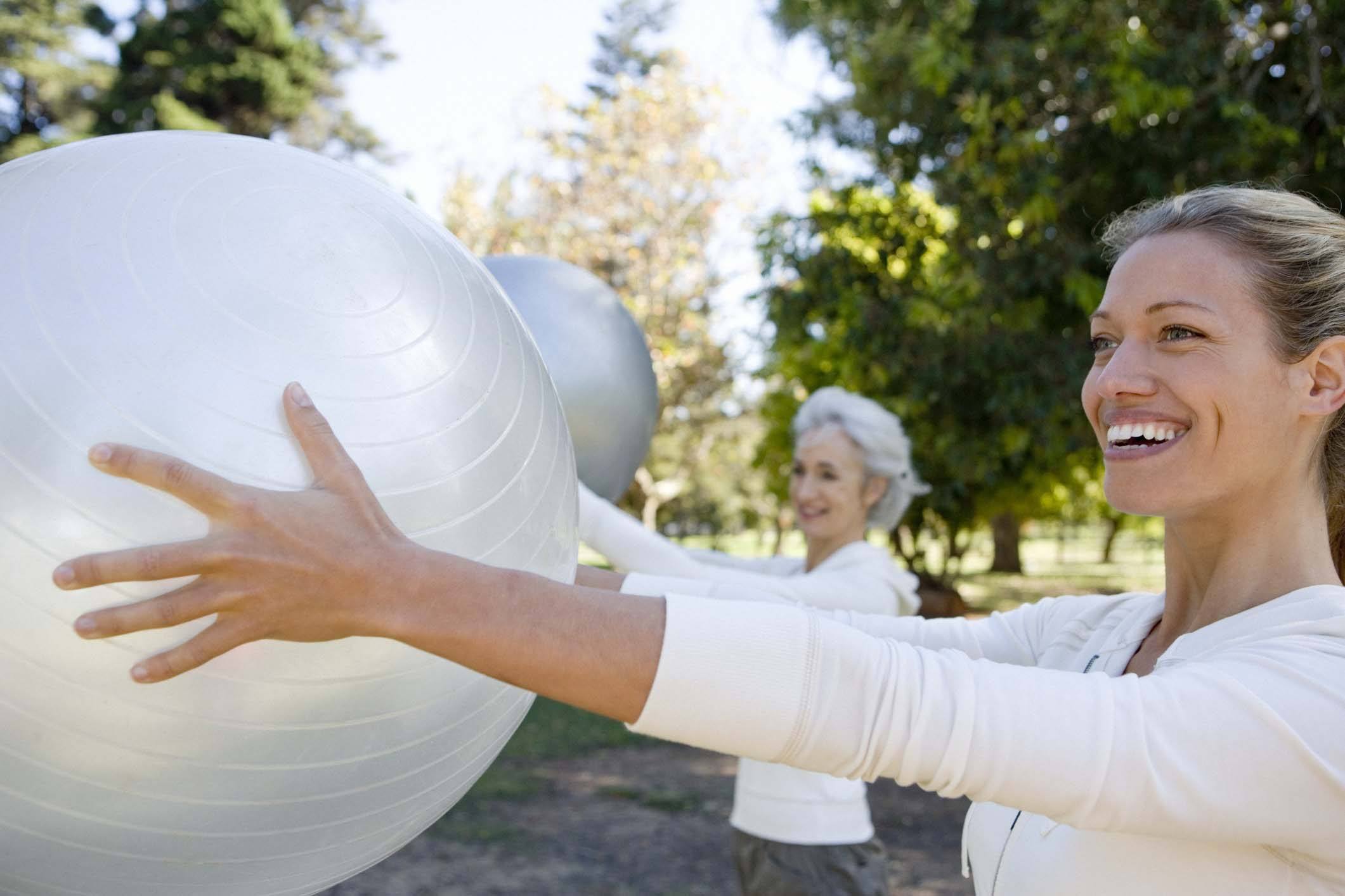 Two women exercising outdoors in a park holding large fitness balls, smiling