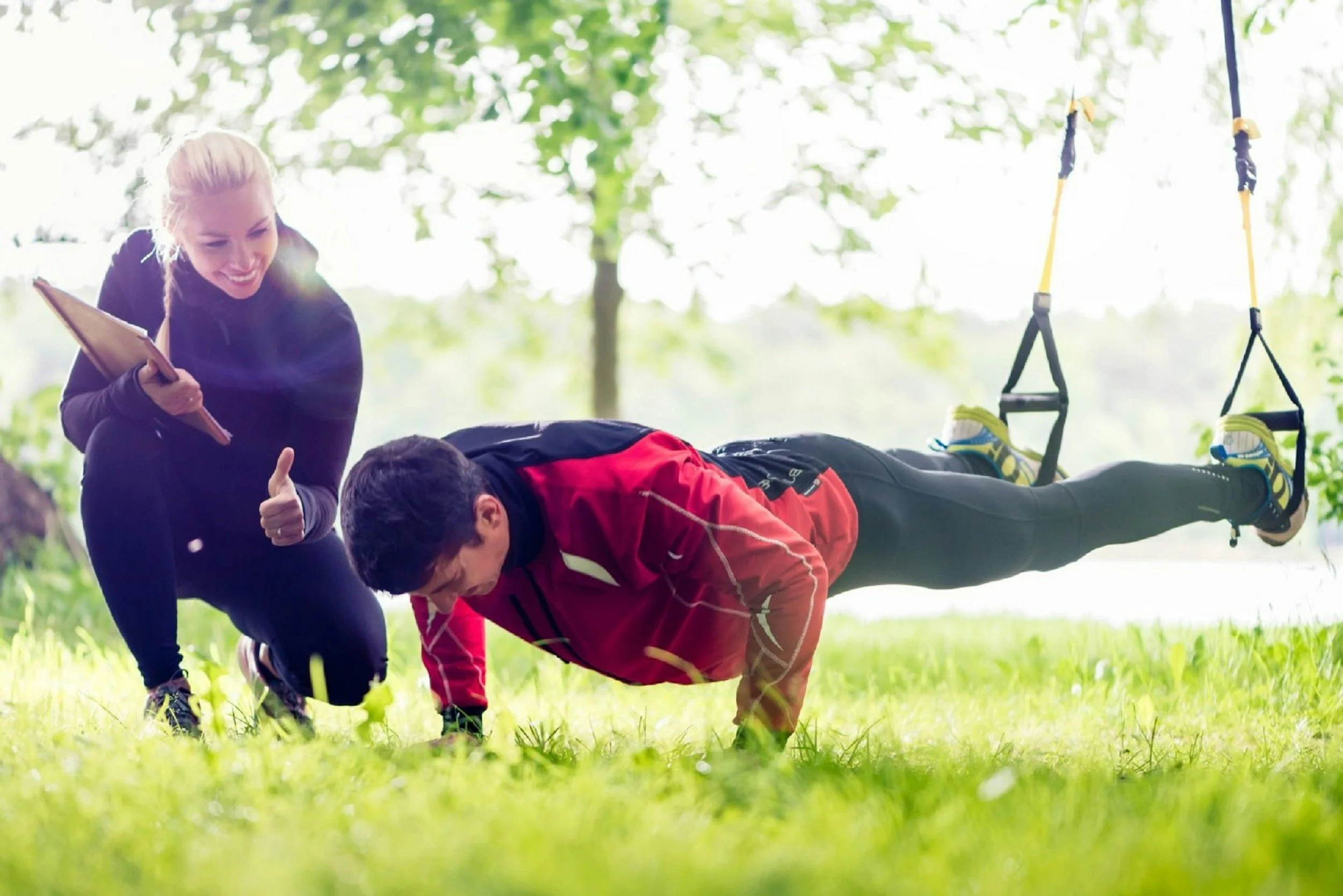 Female trainer coaching a male client doing a TRX plank push-up outdoors on grass