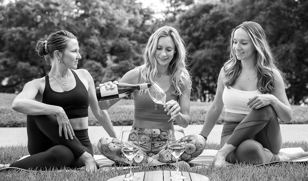 Three women in yoga attire sitting on the grass pouring wine into glasses