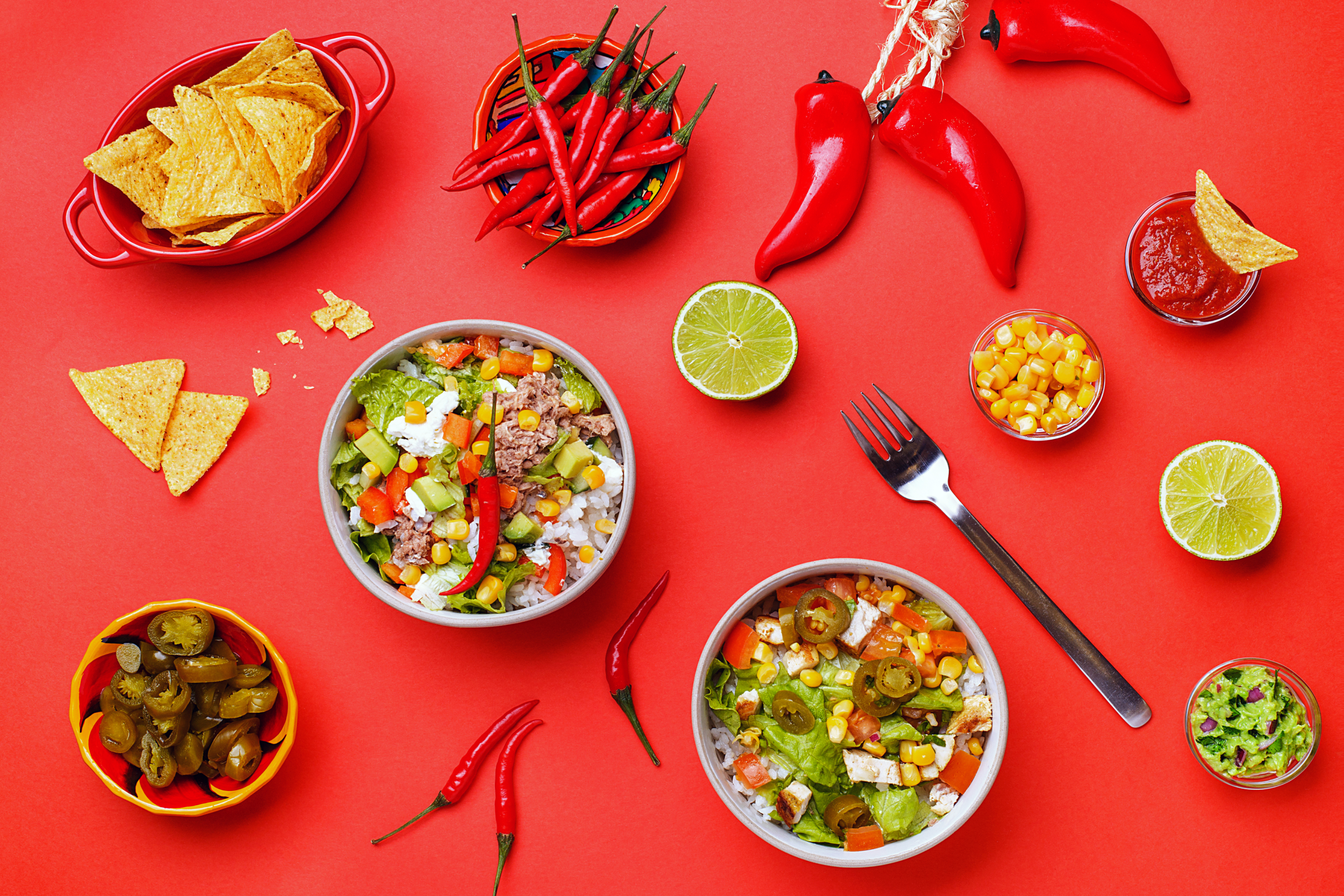 Overhead flat-lay of Mexican bowls, chips, chilis, limes, guacamole and salsa on red